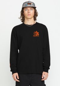 Schwarzes Langarm-T-Shirt mit einem orangefarbenen Logo auf der linken Brust und strukturiertem Ärmel-Detail. Das Modell trägt eine blaue gemusterte Kappe.