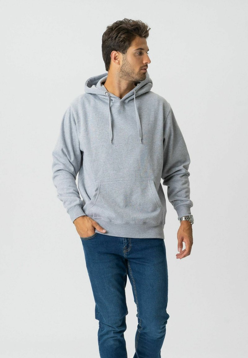 Homme portant un sweat à capuche gris clair et un jean bleu, debout avec une main dans la poche, regardant vers sa droite.