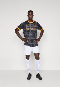 Mandlig fodboldspiller står med hænderne på hoften iført en sort og orange mønstret trøje, hvide shorts, hvide sokker og sorte fodboldstøvler.