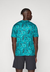 Homme avec des cheveux tressés portant une chemise à manches courtes noire et turquoise à motifs et un short noir, se tenant de dos devant un fond uni.