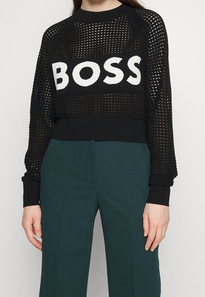 Persoon draagt een zwarte mesh trui met de witte letters "BOSS", gecombineerd met donkergroene, hooggetailleerde broek tegen een lichte achtergrond.