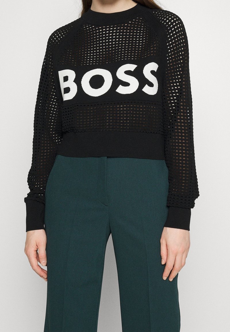 Personne portant un pull en maille noir avec l'inscription "BOSS" en lettres blanches, associé à un pantalon vert foncé taille haute, sur un fond clair.
