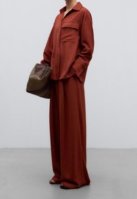 En rustfärgad skjorta med krage och bröstficka, kombinerad med vida byxor. En strukturad brun och beige hatt hålls i handen.