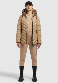 Veste matelassée beige avec capuche, comportant des motifs de piqûres en zigzag et des cordons de serrage. Associée à un ensemble beige assorti et des bottes noires jusqu'aux genoux.