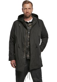 Parka negra con capucha, con cordones, cremallera frontal y bolsillos laterales. Llevada sobre una camisa de camuflaje verde, presenta un corte relajado y una tela duradera.