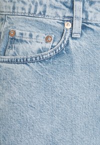 Jeans de mezclilla de color azul claro con una textura deslavada, que cuentan con herrajes de botón en color cobre y un bolsillo frontal. La costura es visible a lo largo de las costuras.