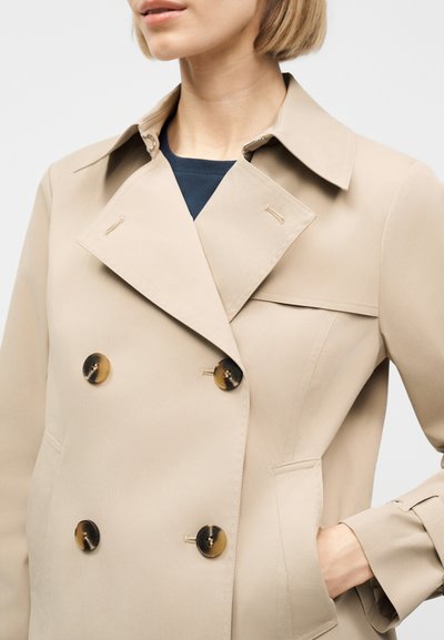 Femme portant un trench-coat beige croisé avec des boutons écaille de tortue, une main dans la poche, et une chemise noire visible au col.