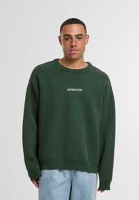 Zeleni pleteni pulover s okruglim izrezom i rebrastim rubom, s natpisom "ANOTHER COTTON" u bijelom na prednjem dijelu. Mekana tekstura.