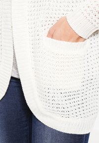 Die Hand ruht in der Tasche eines weißen gestrickten Cardigans, der über blauen Jeans getragen wird.