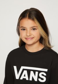 Sort langærmet skjorte med stort hvidt "VANS" logo. Blødt bomuldsstof, rund halsudskæring. Håret er mørkebrunt med lysere højdepunkter.
