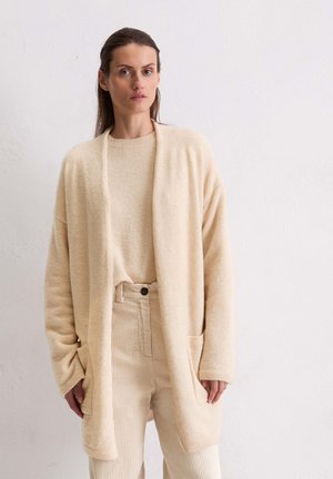 Beige cardigan met een open voorkant, gemaakt van zachte, gestructureerde stof. Gecombineerd met een bijpassende trui en corduroybroek, beide in vergelijkbare kleuren.