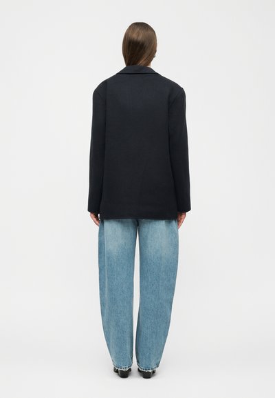 Oseba z dolgimi lasmi, oblečena v črni blazer in široke svetlo modre kavbojke, stoji obrnjena stran proti belemu neizrazitemu ozadju.