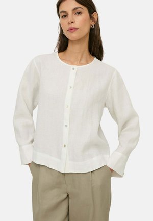 Mujer con blusa blanca de manga larga abotonada y pantalones beige, de pie con las manos en los bolsillos frente a un fondo liso.