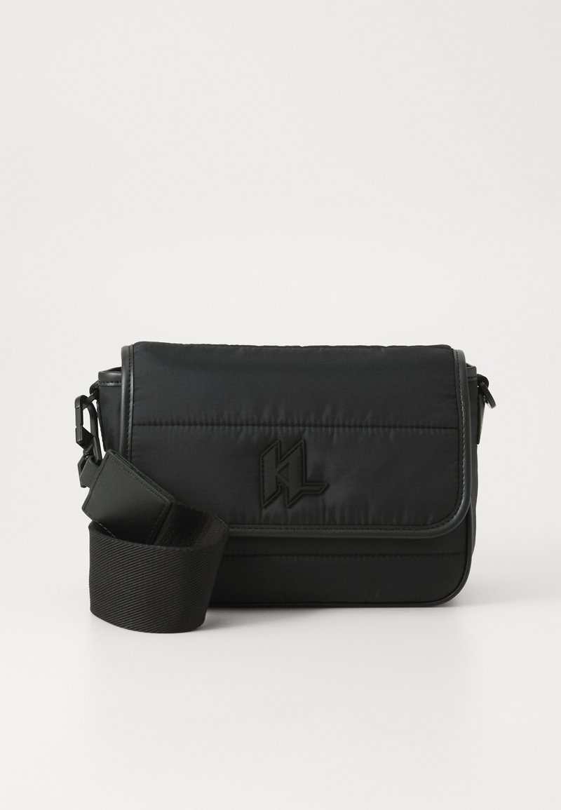 Sac messager noir en nylon, avec un design matelassé, une sangle réglable et un logo embossé à l'avant. Texture lisse.