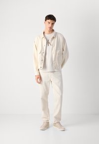 Young man standing, wearing off-white jacket, Calvin Klein logo T-shirt, matching pants, and beige sneakers, against a plain white background.
