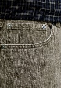 Gros plan sur la poche d’un pantalon en tissu gris texturé avec rivets métalliques, associée à une chemise bleu foncé à motif quadrillé blanc.