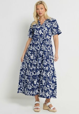 Vrouw draagt een midi-jurk met blauwe en witte bloemenprint, korte mouwen en witte sleehakken, tegen een effen achtergrond.