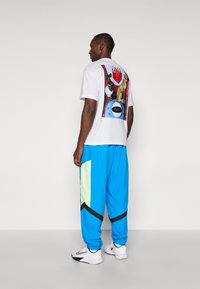 White T-shirt with a colorful graphic on the back, paired with blue track pants featuring mesh accents and black angular details. White sneakers.