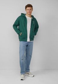 Grüne Zip-Hoodie mit Kängurutasche, getragen über einem weißen T-Shirt. Kombiniert mit hellblauen, verwaschenen Jeans und hellen Turnschuhen.