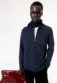 Navy pullover with long sleeves, half-zip neckline, chest pocket, and embroidered accent. Textured scarf wrapped around the neck.