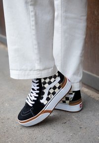 Zapatillas altas en blanco y negro con diseño de cuadro, hechas de ante y lona, con cordones blancos y una suela de goma con acentos marrones.