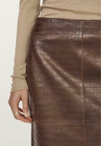 Jupe en cuir marron à motif crocodile avec une texture lisse, affichant une coupe près du corps et un éclat subtil, tenue par une main portant un haut à manches longues beige.