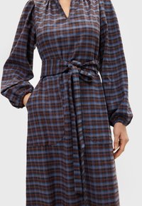 Brun og blå rutete kjole med V-hals, puffede ermer og knyttet midjebånd. Har to sidelommer og myk tekstur.