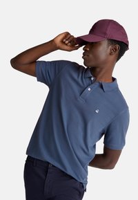 Mladý muž v modrém polo tričku a vínovém klobouku, naklání hlavu dolů a jednou rukou si upravuje kšilt klobouku.