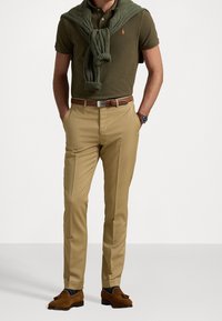 Mann trägt ein olivgrünes Polo, einen passenden Pullover lässig über die Schultern gelegt, beigefarbene Chinos, einen braunen Gürtel, eine Uhr und braune Quastenloafer mit dunklen Socken.