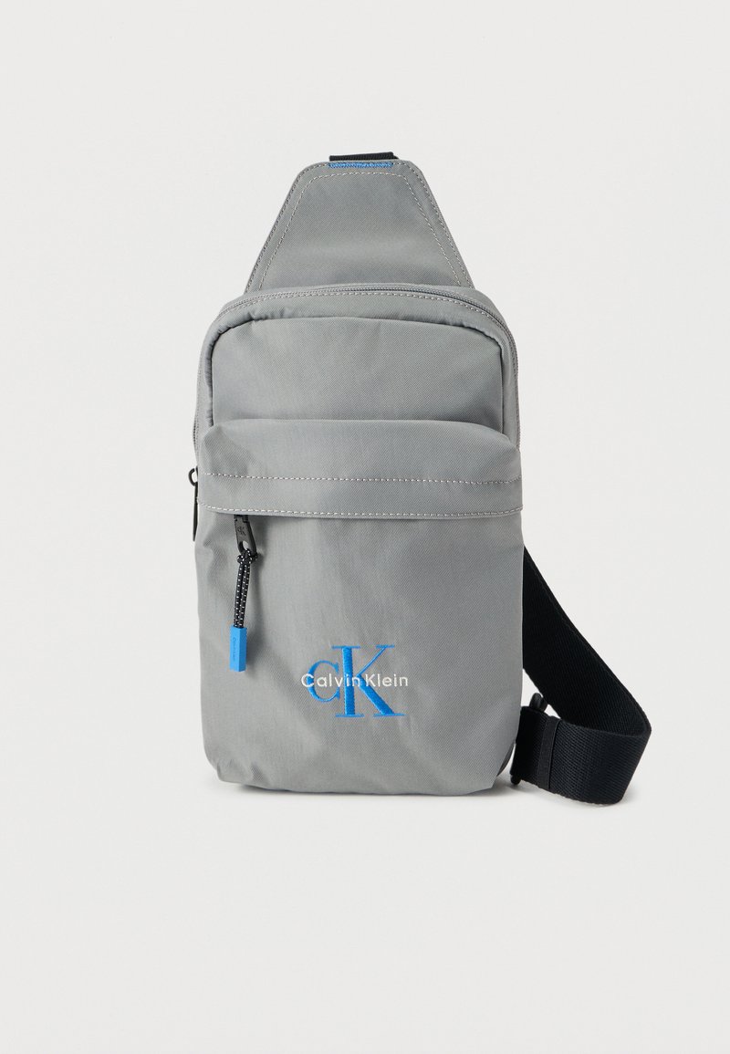 Γκρι τσάντα ώμου από νάιλον με μπροστινή τσέπη με φερμουάρ, μπλε λογότυπο, ρυθμιζόμενο ιμάντα και απαλό υφή με μινιμαλιστικό σχεδιασμό.