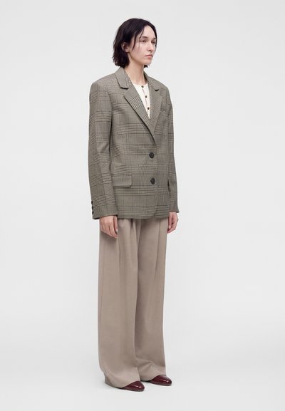 Femme debout portant un blazer à carreaux oversize, un top blanc à boutons, un pantalon large beige et des chaussures pointues foncées sur un fond uni.