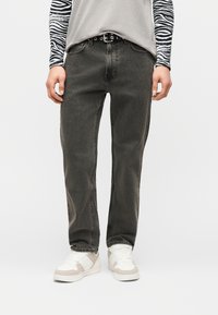 Άτομο που φοράει ξεθωριασμένο μαύρο τζιν με ευθείες μπότες, λευκά και μπεζ αθλητικά παπούτσια, μακρυμάνικα με σχέδιο ζέβρας κάτω από γκρι μπλουζάκι, όρθιο.