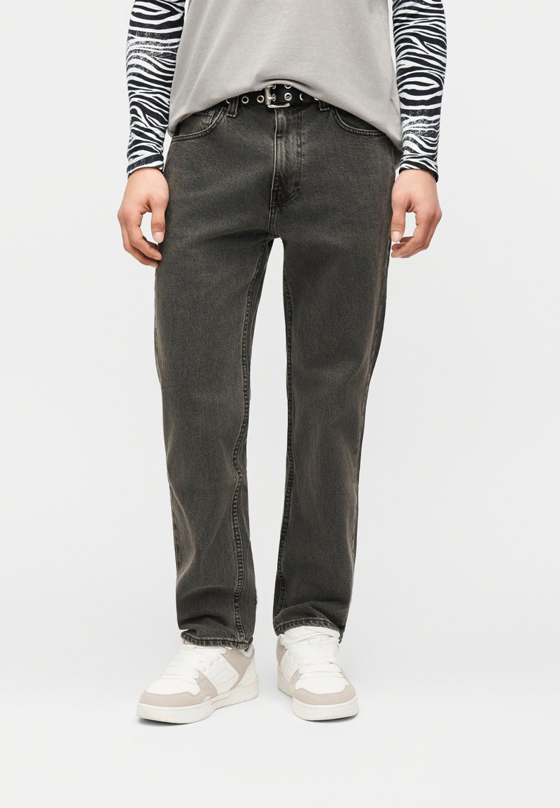 Άτομο που φοράει ξεθωριασμένο μαύρο τζιν με ευθείες μπότες, λευκά και μπεζ αθλητικά παπούτσια, μακρυμάνικα με σχέδιο ζέβρας κάτω από γκρι μπλουζάκι, όρθιο.