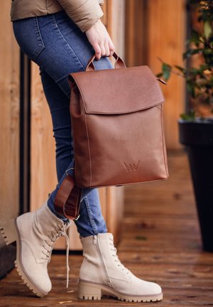 Hnědý batoh z ekologické kůže s klopou, dvojitými uchy a nastavitelným ramenním popruhem, který na přední straně má jemně embosovanou logo.