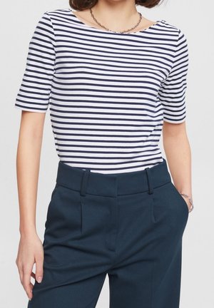 Vrouw draagt een wit shirt met korte mouwen met marineblauwe horizontale strepen en marineblauwe hoog getailleerde broek met handen in de zakken.