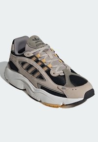 Sneaker with a beige and black mesh upper, grey suede accents, yellow laces, and a thick white outsole. Features three black stripes.