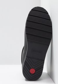 Semelle de botte en caoutchouc noir avec un profil texturé pour l'adhérence, un logo circulaire rouge et le nom de la marque embossé dans le matériau.