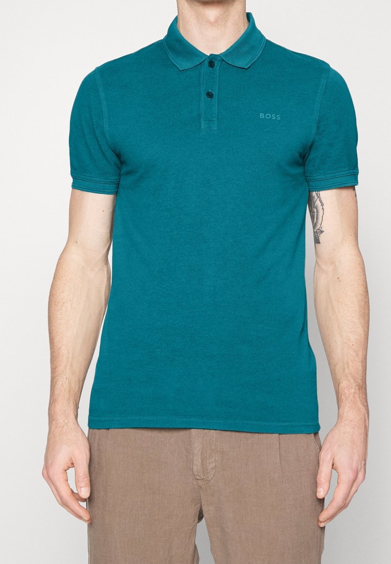 Polo tee bleu-vert en coton doux, avec un col classique, une patte de boutonnage à deux boutons et un détail logo subtil sur la poitrine. Manches courtes.