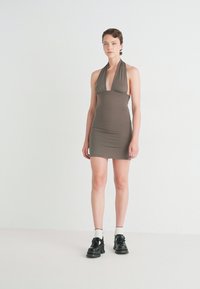 Halter-neck, nøytral brun kjole med en figursydd design. Mykt stoff med minimal strekk. Kombinert med svarte sko og hvite sokker.