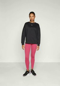 Mujer de pie con un sudadera negra de Puma de manga larga, leggings rosas y zapatillas deportivas negras contra un fondo liso.