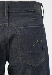 G-Star Raw tamnoplave traperice s kontrastnim žutim šavovima, prepoznatljivim dizajnom stražnjeg džepa i brendiranim kožnim oznakom na struku.