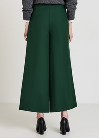 Pantaloni a gamba larga in tessuto verde scuro, con una texture liscia e un taglio sartoriale, abbinati a scarpe nere con tacco spesso.