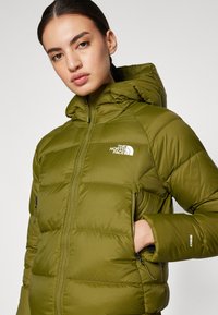 Maslinasto zelena puffer jakna s kapuljačom, zatvorenim džepovima i kviltanim dizajnom. Ima bijeli logo na prsima i oznaku 850 na rukavu.