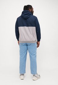 Chaqueta con capucha en azul marino y gris claro con un diseño de bloques de color, puños elásticos y dobladillo. Lleva puesta con jeans de color azul claro y zapatillas grises.