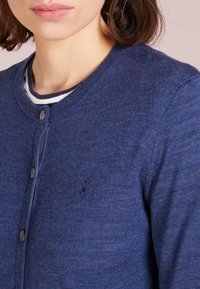 Blå cardigan i texturerad stickad kvalitet, med rund halsringning, knäppning och en liten logotyp på vänster bröst.