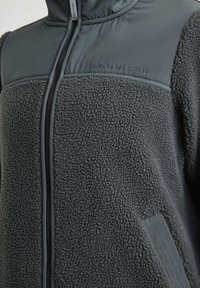 Tummanharmaa fleece-takki, jossa on sileä yläosa ja karheapintaista alaa, korkea kaulus, vetoketju ja kohokuvioitu logo-ysityksessä.