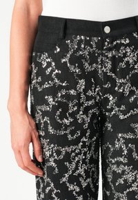 Roka atpūšas blakus melniem biksēm, kuras ir rotātas ar izkaisītām spīdīgām sudraba pūcēm un sudraba pogu jostasvietā.