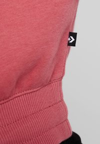 Acercamiento de un puño de tela acanalada rosa con una etiqueta negra que presenta un logo de estrella blanca sobre un fondo gris.