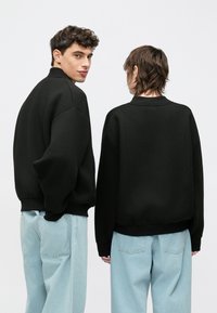 Zwarte oversized sweatshirts met geribbelde kragen, gedragen door twee personen die met de rug naar de camera staan. Lichtblauwe loszittende jeans completeren de outfits.