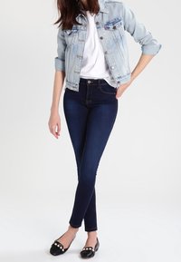 Kvinna i ljusblå denimjacka, vit skjorta, mörka tajta jeans och svarta ballerinas med pärldekor, poserar mot vit bakgrund.